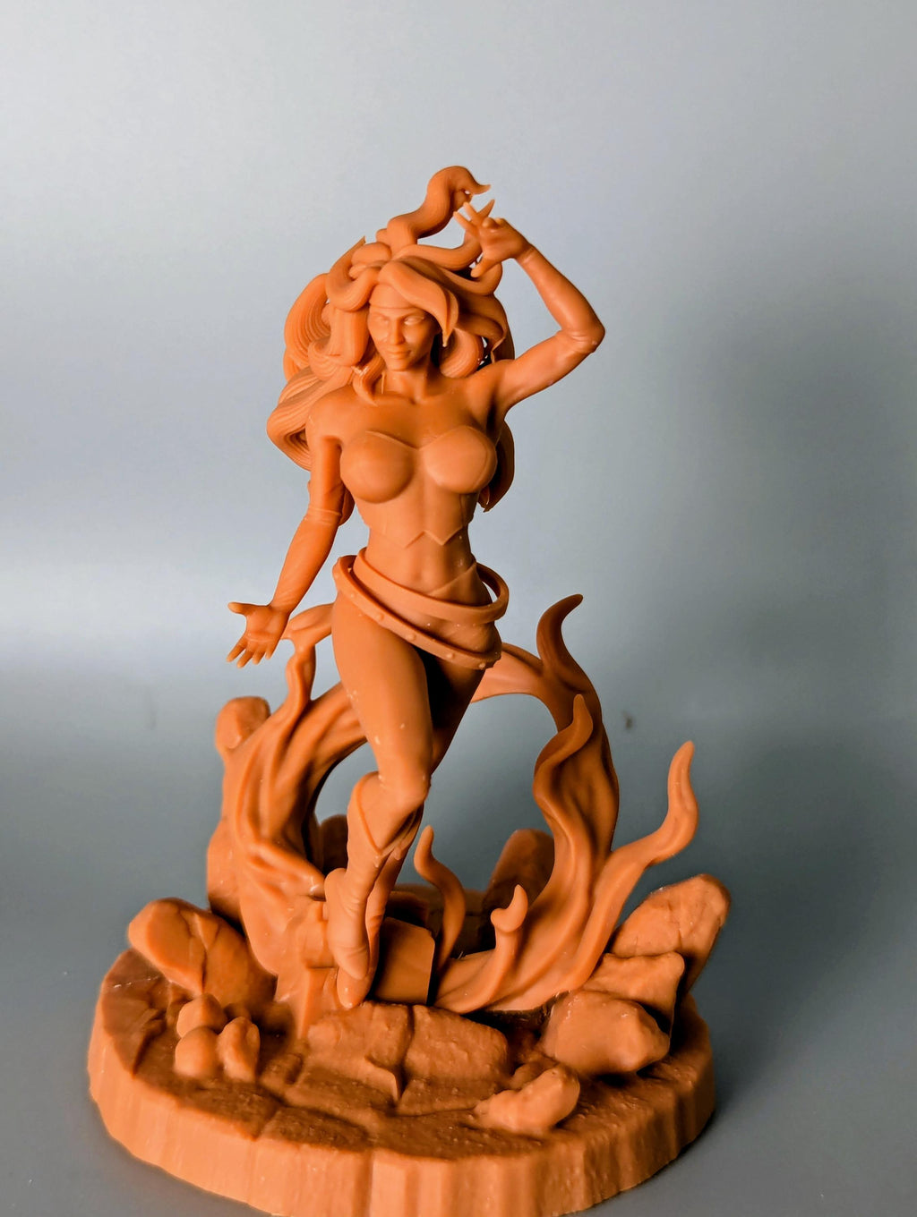 a figurine of a woman with a large head and long hair, standing on a rock formation with flames or fire behind her.