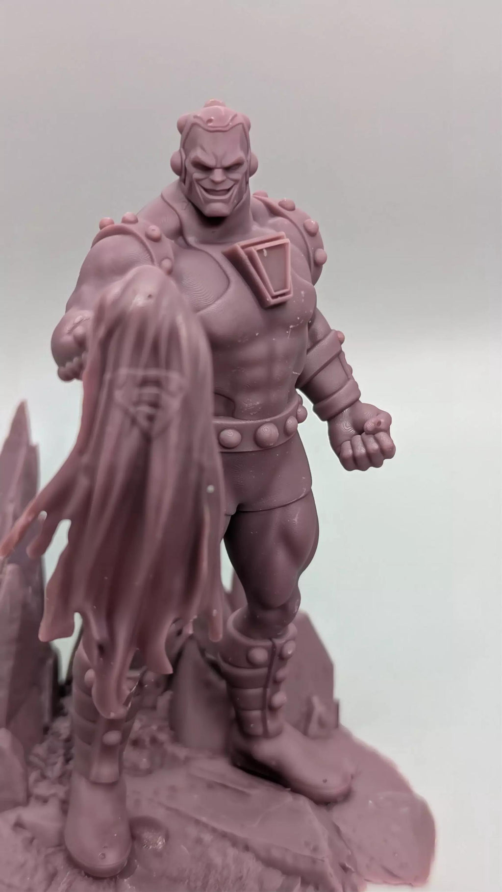 A close-up image of a figurine of a muscular, armored figure with a cape-like design on the back, standing on a rocky surface.