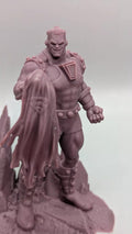 A close-up image of a figurine of a muscular, armored figure with a cape-like design on the back, standing on a rocky surface.