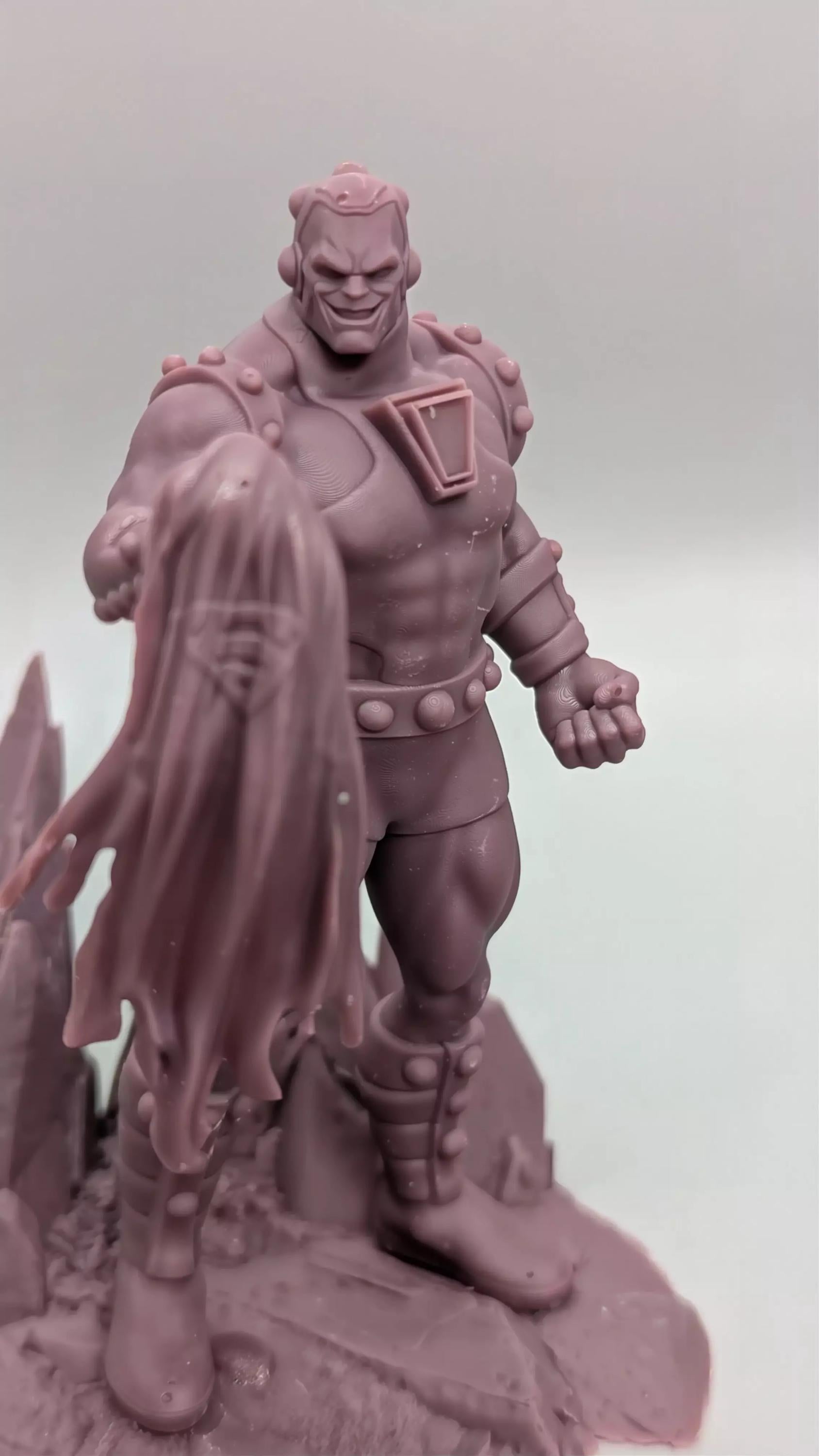 A close-up image of a figurine of a muscular, armored figure with a cape-like design on the back, standing on a rocky surface.